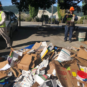 two workers in hard hats and safety vests standing next to a pile of cardboard and paper being sorted for recycling