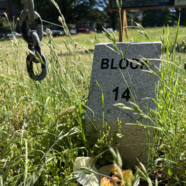 A stone marker with the words "Block 14" engraved in black letters stands on a grassy field with wildflowers in the foreground and an oxidized chain hanging from above