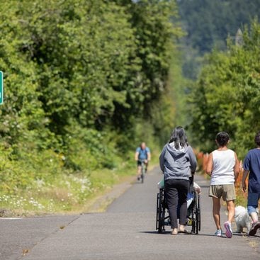 A group of three people walk along the Gresham Fairview Trail. One person pushes another person in a wheelchair. A person on a bike is approaching from further down the trail. Trees line both sides of the trail on a sunny day.