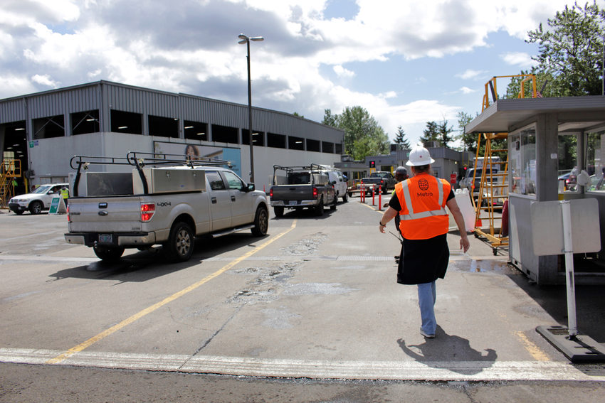 A worker wearing a reflective vest and hardhat walks near a line of trucks waiting to enter a large industrial building