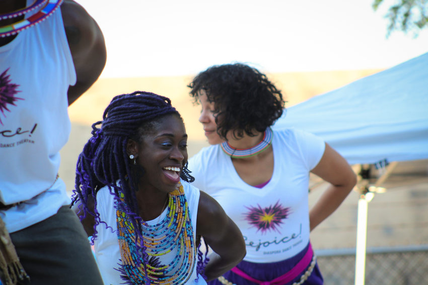 Dancers from Rejoice Diaspora Dance Theater performing on stage. The middle dancer smiles wide as they all lean forward with their hands behind their back. They each wear white shirts with colorful logos