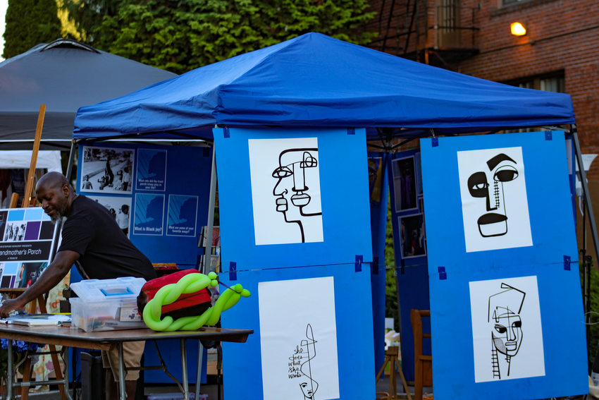 A Black man stands in front of a blue tent that has art pieces displayed on its side