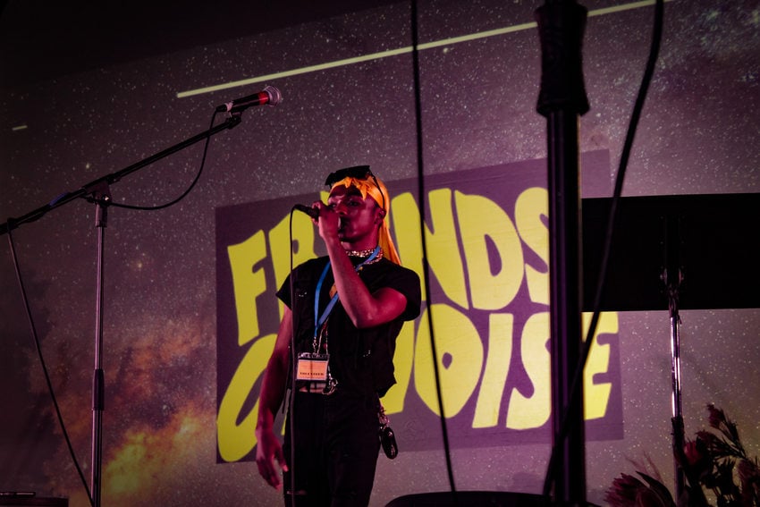 A youth by the name Daboi King raps on stage at the Afro-topia event. The Friends of Noise logo is on the projector in the background