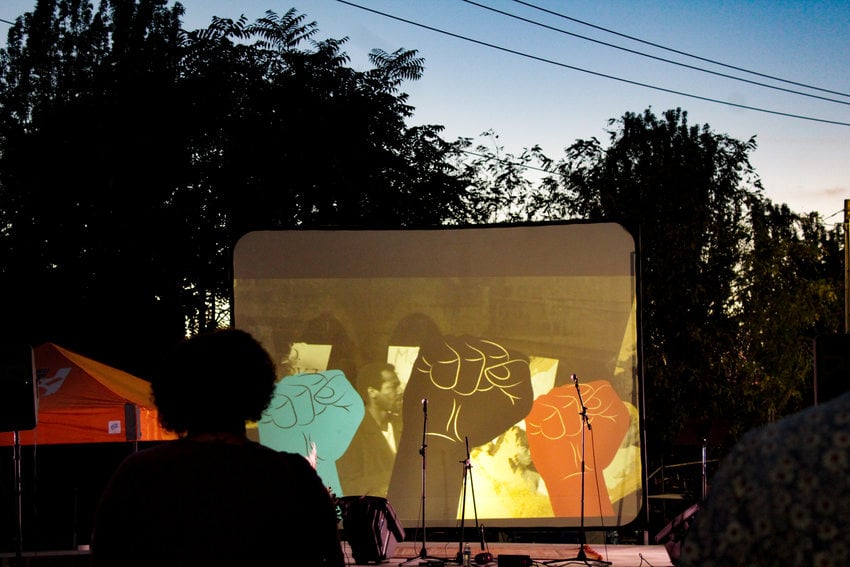 A large projector screen shows a short film and the freeze frame shows multiple fists held up in the air