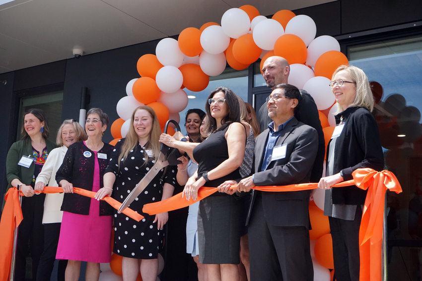 Eleven people gathered under a balloon arch, cutting an orange ribbon with giant scissors to celebrate the opening of an apartment building.