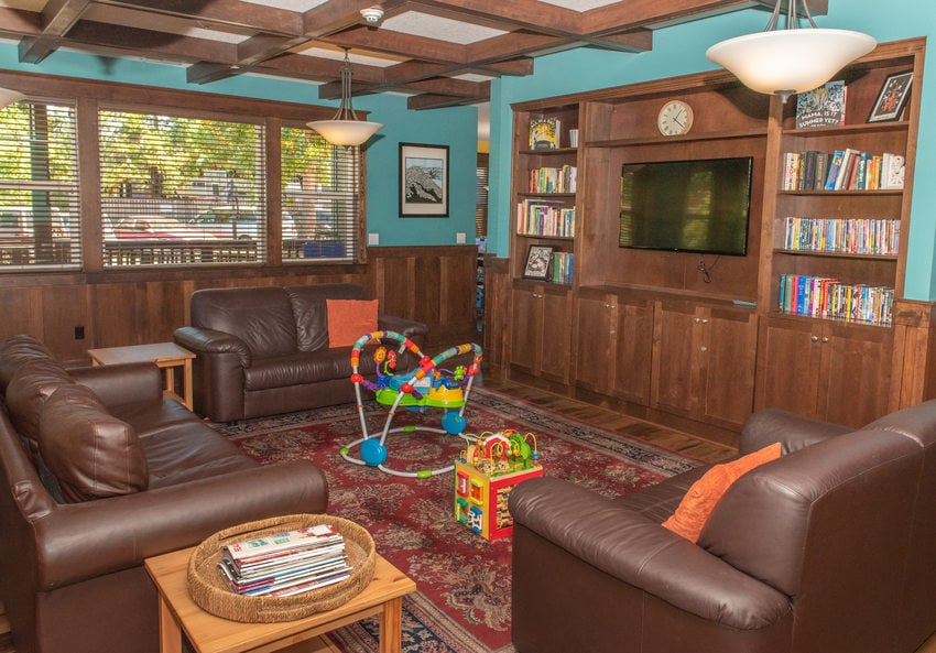 A cozy living room featuring two brown leather sofas and a matching armchair arranged around a central wooden coffee table with magazines on top. There's an entertainment unit on the far wall housing a large TV and shelves filled with books. 