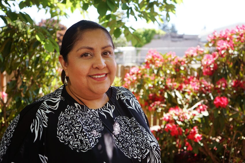 Woman in floral print blouse standing in front of flowering shrubbery and smiling at the camera.