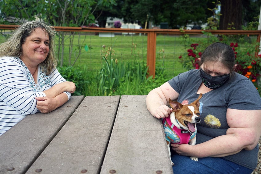 Two women at a picnic table in a grassy setting. One is looking at the camera and the other is looking at her small dog, which is sitting in her lap. 