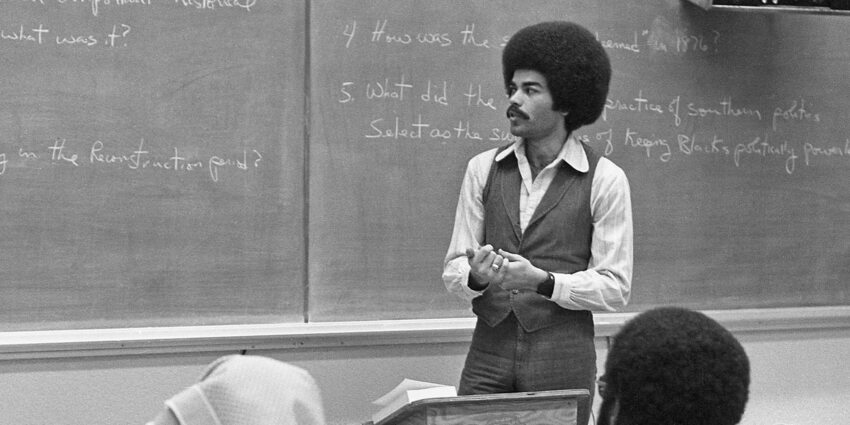 A man with an afro hairstyle stands in front of a chalkboard, gesturing as he speaks to a seated audience. The chalkboard contains handwritten notes and questions.