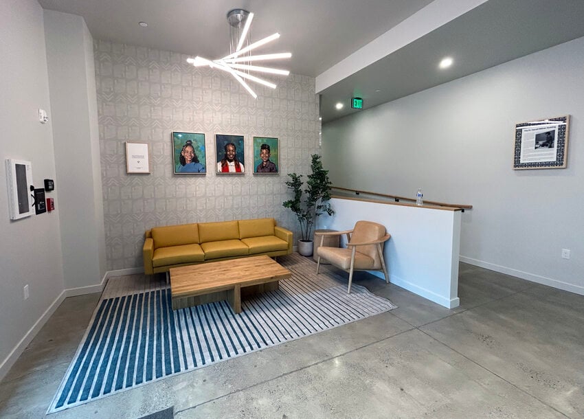 A modern waiting area with a yellow sofa, wooden table, and a beige armchair on a striped rug. Three framed portraits are hung on a textured wall. A potted plant sits in the corner.