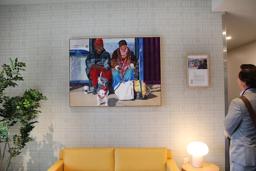 A person in a grey suit looks at a painting of two people at a bus stop. The painting is displayed in an apartment lobby over a yellow sofa.