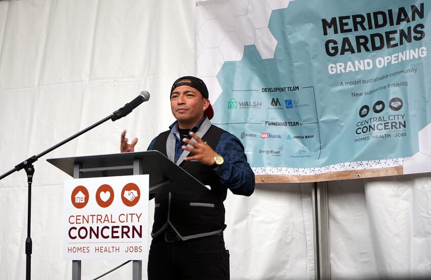 Man wearing a vest and backwards baseball cap speaks at a podium. Signage reads "Meridian Gardens grand opening" and "Central City Concern."