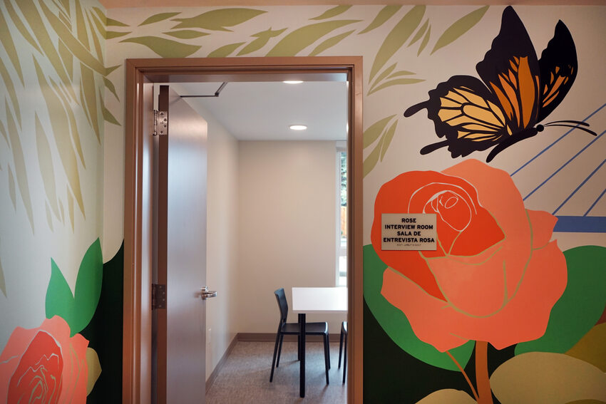 A decorative door featuring a colorful butterfly perched on a rose. Through the doorway is a table and chairs with a large window. The sign by the door reads "Rose Interview Room."