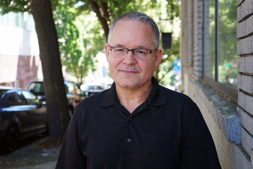 Portrait of a middle-aged man with black collered shirt and glasses, standing on a tree-lined downtown street. 