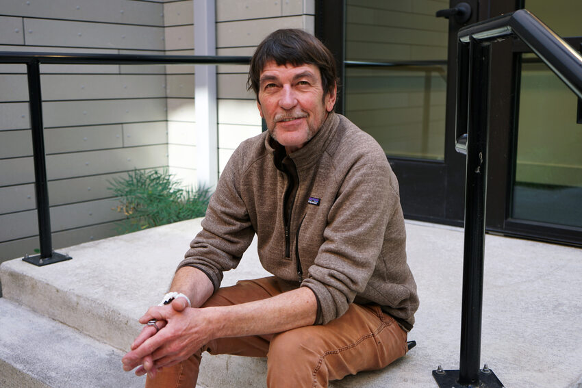 Middle aged man siting on the steps in an apartment building courtyard. He's wearing a brown sweatshirt and brown pants.