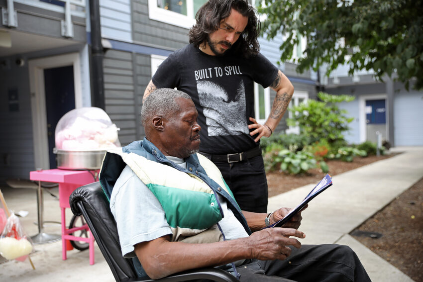 A young man and an older man in the courtyard of an apartment building. They are looking at a document together and there is a cotton candy machine in the background.