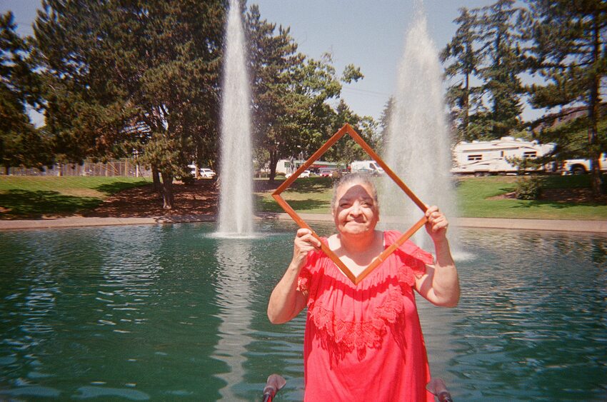 Older woman in a red shirt posing in front of a fountain, holding a picture frame in front of her face.