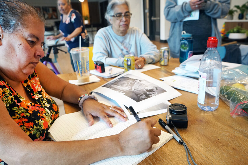 Woman sitting at a table with other women, writing in a notebook. 