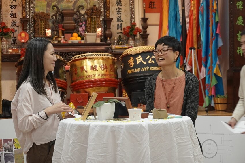 Two women stand, smiling at each other, at a tall round table with Chinese drums in the background