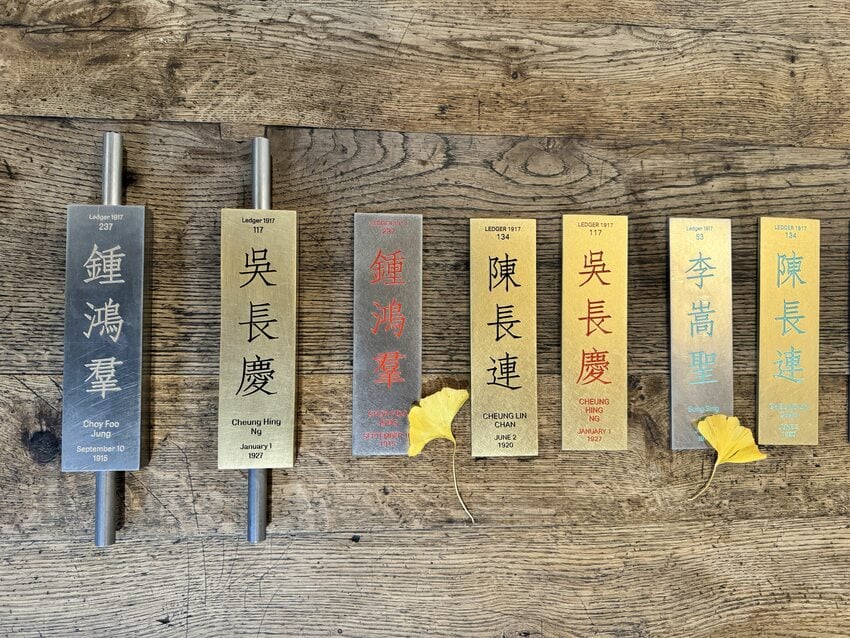 a line of seven metal rectangular tiles inscribed with Chinese and English writing sit on a wooden table with gingko leaves scattered about