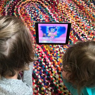 two little kids listen to an audio book on a tablet while laying down on a rug on the floor