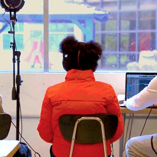 A group of young people sitting in a classroom with headphones on in front of a laptop. A microphone on a stand is nearby.