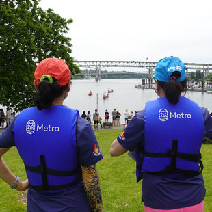 Two women seen from behind wearing Metro life jackets on Poets Beach, watching dragon boats approach on the river