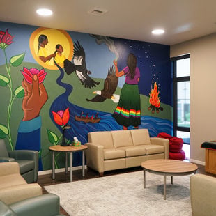 A community room with a large mural depicting various scenes: an elder and child surrounded by flowers, an eagle in flight, and a group in a canoe on a river.