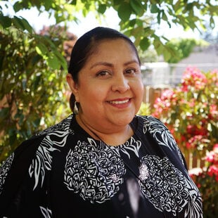 Woman in floral print blouse standing in front of flowering shrubbery and smiling at the camera.