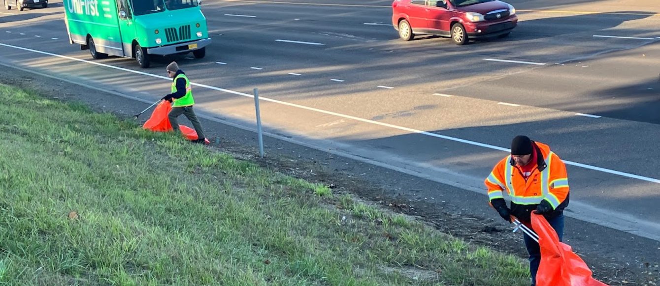 A patch of grass on the side of the highway being cleaned by two people in high visibility gear. Cars pass by in the background.
