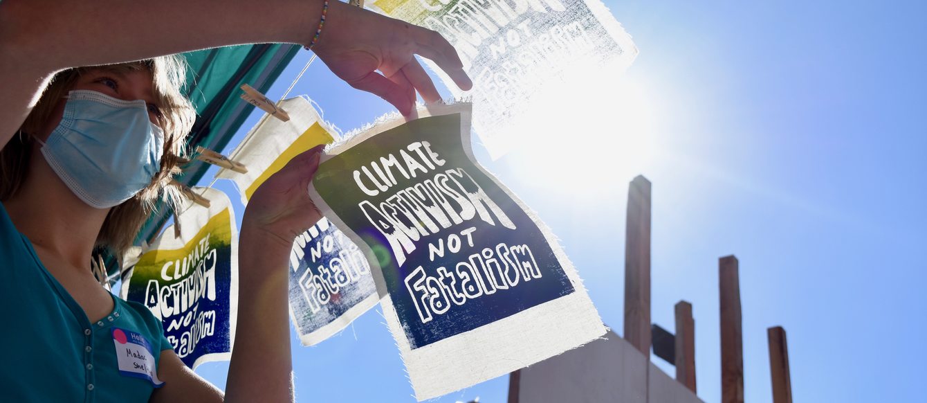 A young climate activist gets ready to hang a sign up to dry. The signs have a blue and yellow background and say "Climate Activism Not Fatalism."  The sun shines bright in the clear blue sky, and a factory looms in the backround.