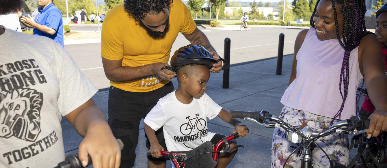 Image of an adult in a yellow t-shirt putting a bike helmet on a child who is sitting on a bike. A woman in a pink top looks at them and smiles