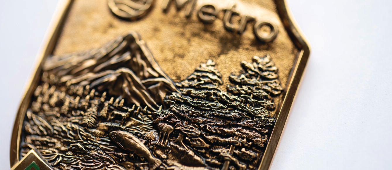 A close up of a bronze park ranger badge has details of mountains, trees and a river. It reads "Metro Park Ranger". 