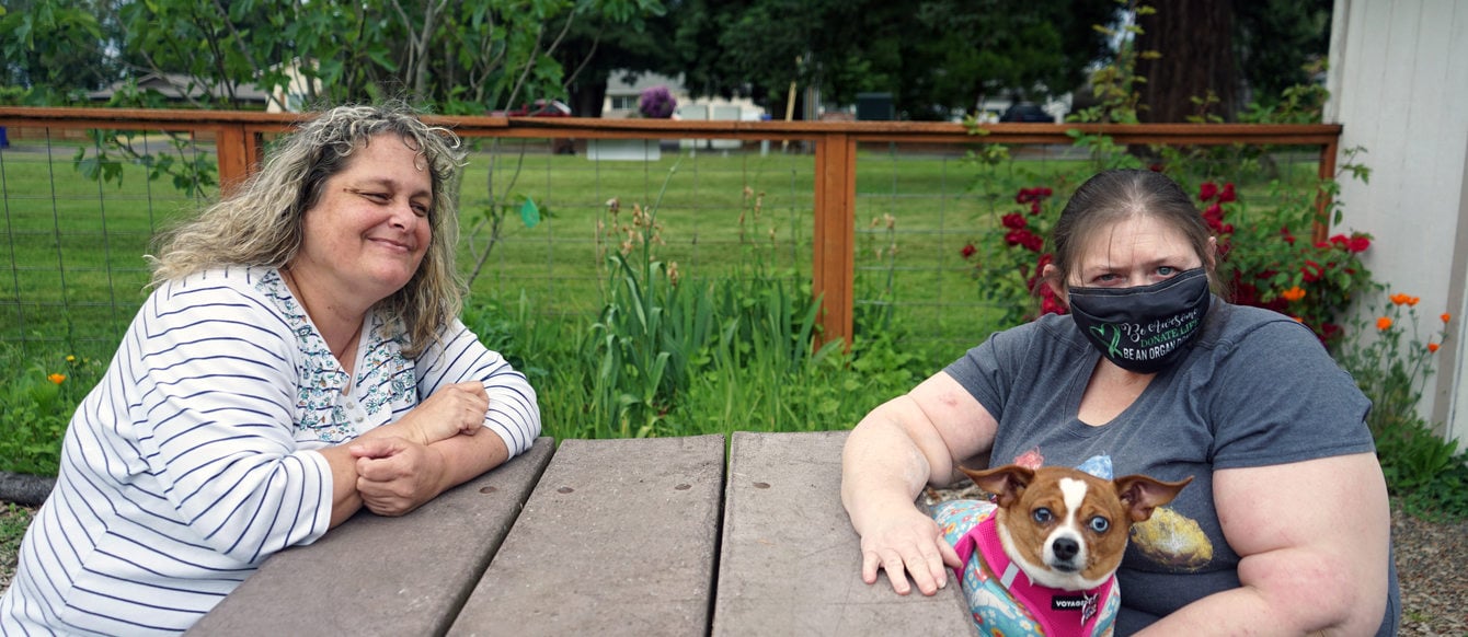 Two women at a picnic table in a grassy setting. One is smiling at the other, who is holding a small dog in her lap.