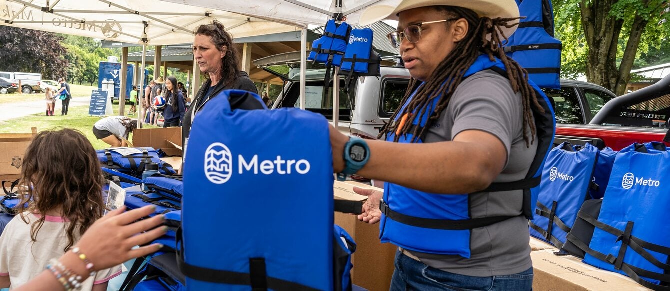 A Metro staff person passes a Metro-logoed life jacket to an outstretched hand at an outdoor festival booth, rows of life jackets stacked behind them