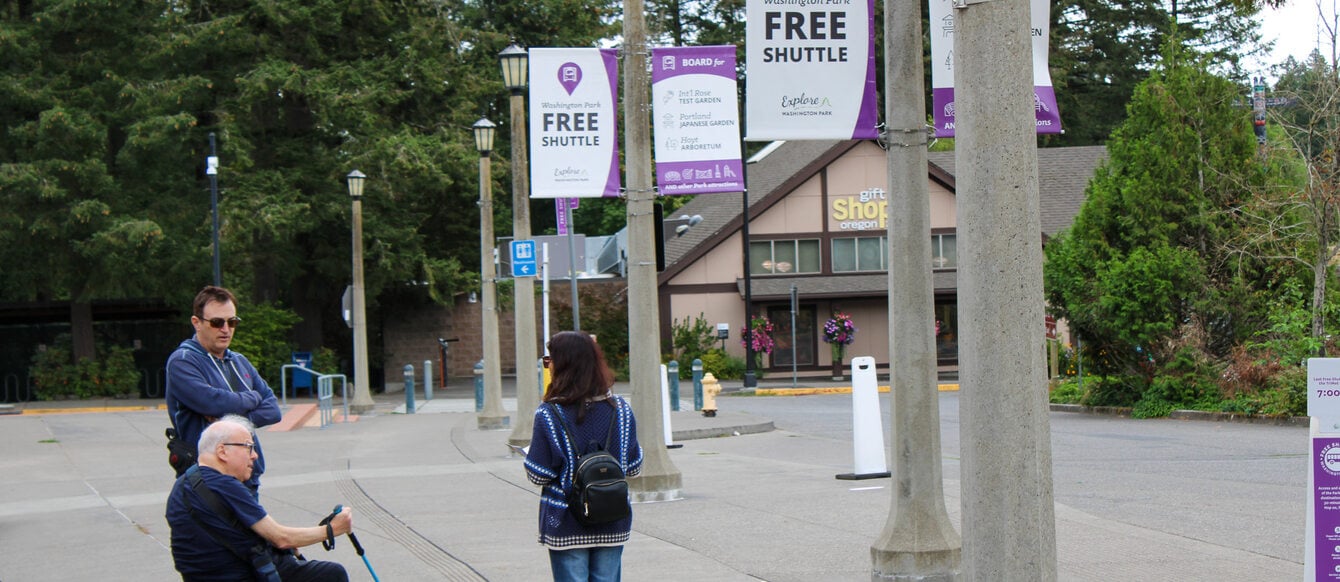 Three people, standing and sitting, wait on the sidewalk for the Washington Park free shuttle. Signs for the free shuttle hang from light posts that line the sidewalk and trees surround the area.