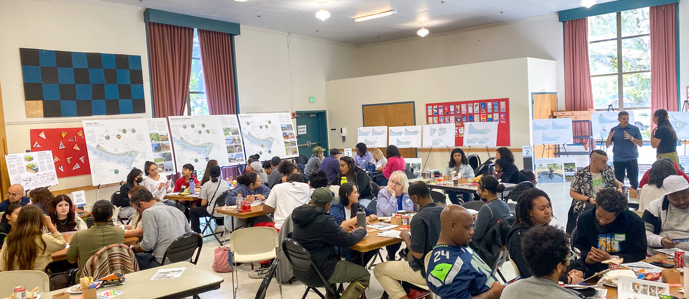 A community center room is filled with about 50 people sitting around folding tables.