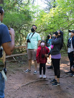 A group of hikers wearing masks listen to a nature educator wearing a large backpack.