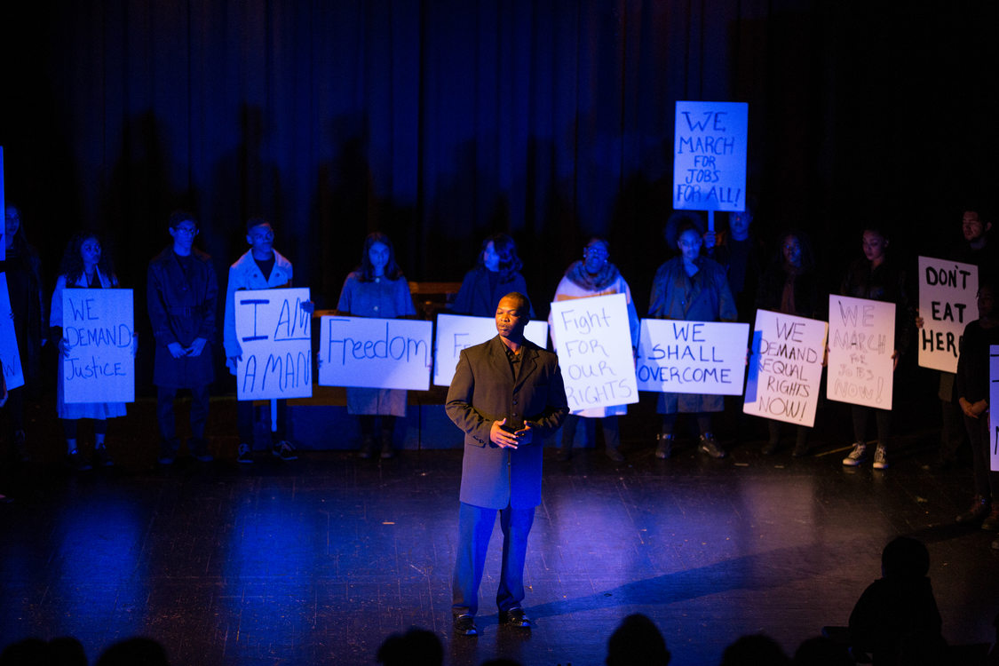 Actors on stage recreating the March on Washington and Dr. Martin Luther King, Jr.'s "I have a dream speech."  Actors hold signs that say "freedom" "we shall overcome" "fight for our rights" and more.