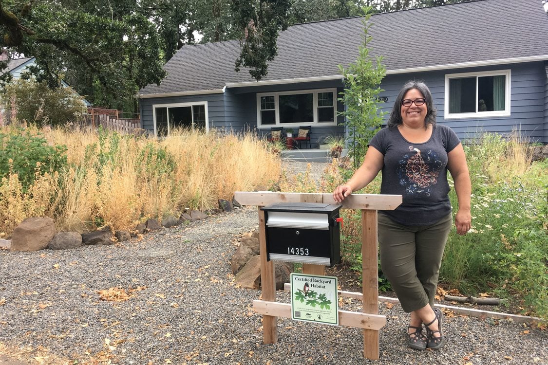 A person stands in front of their home, smiling, their backyard habitat certification sign is displayed under their mailbox and native plant habitat is visible in their front yard.
