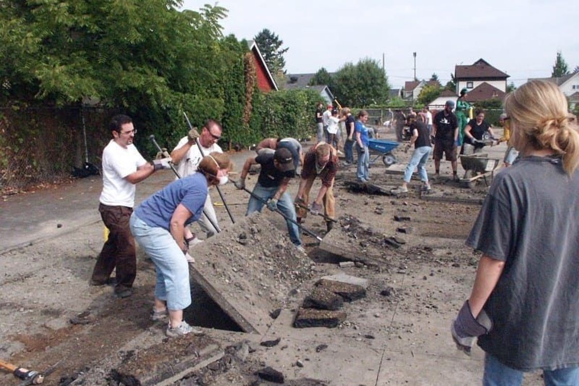 Several adults weilding crowbars or shovels work together to lift large panels of concrete. In the background are more volunteers some doing the same, others loading wheelbarrows and several homes.