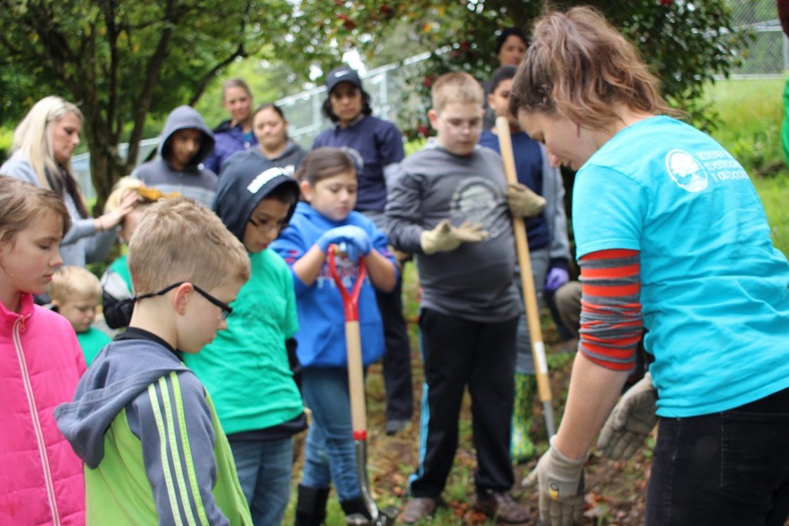 Several elementary age students watch closely, shovels in hand, as a nature educator points their attention to the ground. There are trees and adult observers in the background.