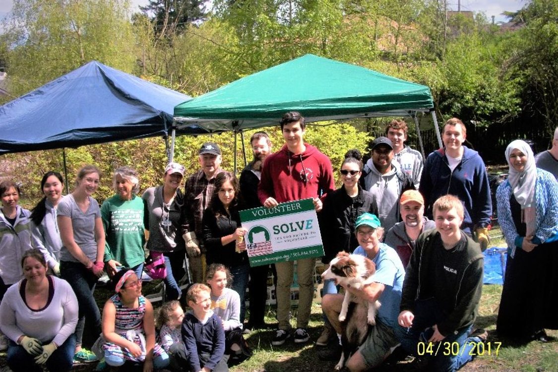 A large group of people ranging in age pose and smile holding a 'Volunteer with SOLVE' sign. There are pop up tents and trees in the background.