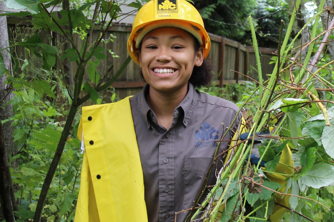 A youth wearing NW Youth Corps shirt, raincoat and hardhat smiles widely holding a pile of cut Himilayan Blackberry in their gloved hands. Greenery and a wood fence are visible in the background.