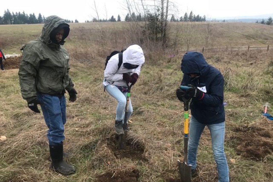 Three people are in a large open field with trees in the background wearing coats with their hoods up. Two youth are using shovels to dig holes for planting.