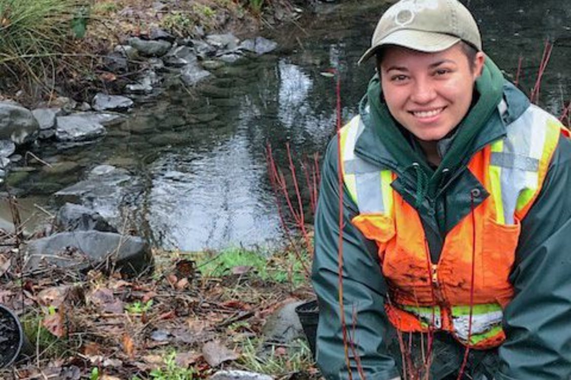 A person wearing rain gear, work gloves and an orange safety vest kneels smiling in front a new planting, a small shovel at their side. Behind them is a calm waterway.