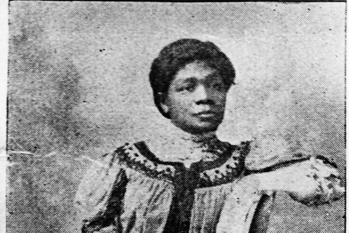 A sophisticated, lovely black woman wearing an embroidered dress and white gloves  poses for a black-and-white photograph
