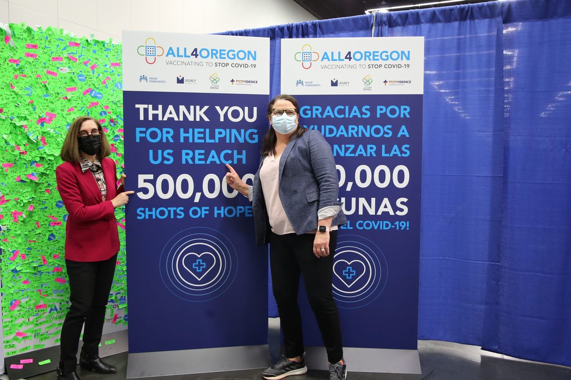 Governor Kate Brown and another woman wear masks and stand next to a banner for All4Oregon at the Oregon Convention Center mass COVID-19 vaccination site