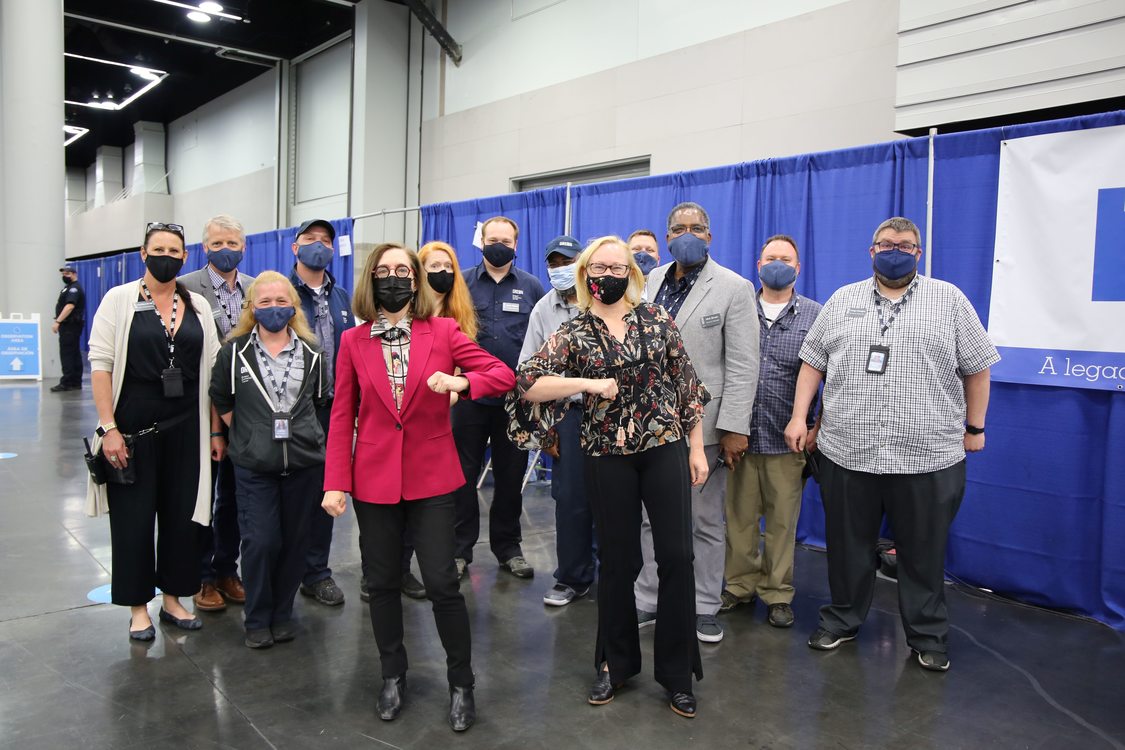 Governor Kate Brown and Metro Council President Lynn Peterson wear masks and stand with a crowd of masked staff and volunteers at the Oregon Convention Center mass COVID-19 vaccination site