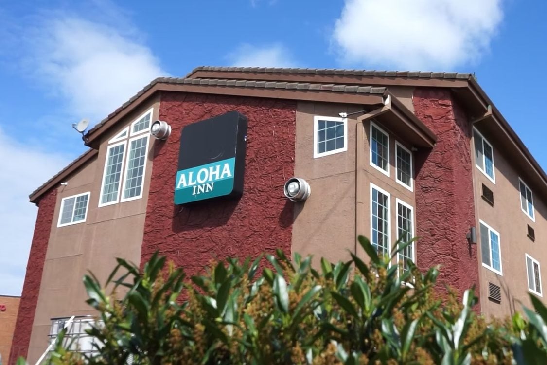 Aloha Inn affordable housing project in Washington County, showing the 3-story building in shades of brown and beige with a teal-colored sign reading Aloha Inn on the side of the building